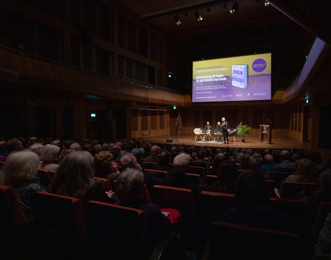 long view of audience facing the stage, with a large digital screen with an image of the One Dublin One Book promotional poster